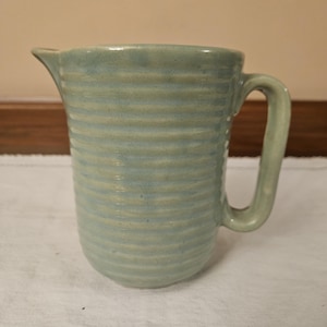 May include: A pale blue ceramic jug with a handle and a pouring spout. The jug features horizontal ridges around its body. It rests on a white cloth with floral embroidery.