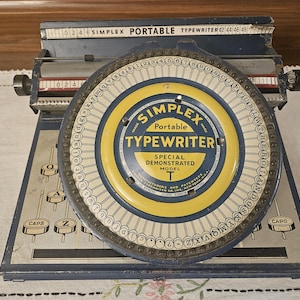 May include: A vintage Simplex Portable Typewriter, Special Demonstrated Model T, in blue and gray. The circular dial features yellow and blue accents with the brand name. The typewriter has a series of keys and a numerical scale.