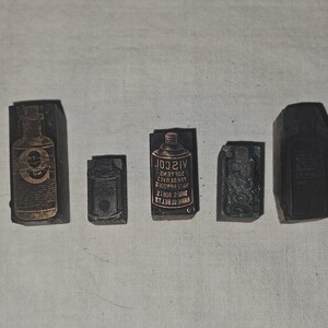 May include: Five vintage printing blocks in a row. The blocks are dark brown and rectangular. The blocks depict bottles and containers, with some text visible on the blocks. The blocks are arranged on a white surface.