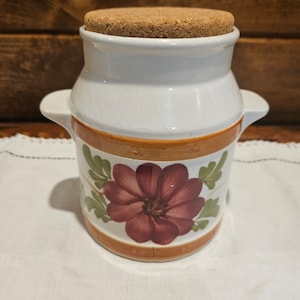 May include: White ceramic jar with a cork lid and a floral design. The jar has two handles and a brown band with a red flower and green leaves.