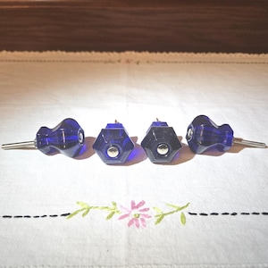 May include: Four cobalt blue glass cabinet knobs with silver metal accents. The knobs have a hexagonal shape and a small screw on the back.