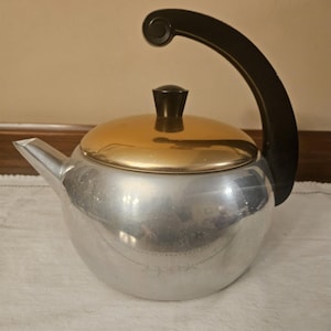 May include: A vintage silver-colored kettle with a gold-toned lid and a black handle. The kettle has a spout and a black knob on the lid. The handle curves over the top of the kettle. The kettle is sitting on a white cloth.