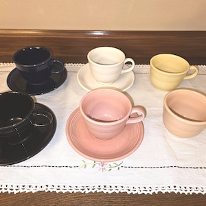 Peut inclure: Six tasses à thé et soucoupes de différentes couleurs, dont noir, blanc, jaune, rose et bleu. Les tasses et soucoupes sont disposées sur une nappe blanche avec une bordure en dentelle.