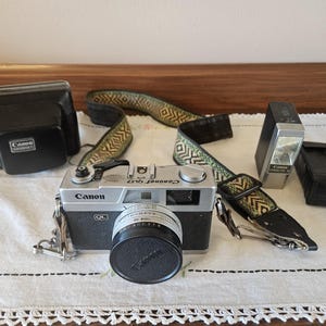 May include: A vintage Canon QL17 camera with a lens cap, a patterned camera strap, a flash, and a black camera case. The camera is silver and black, and the strap has a green and tan geometric design. A black case is also present.