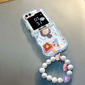 May include: A blue and white phone case with a cartoon bear, cat, and cloud design. The case has a clear back and a white pearl bracelet attached to it. The phone screen shows the time 1:50.