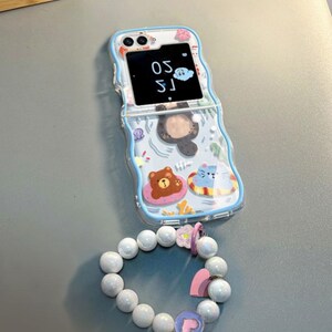 May include: A blue and white phone case with a cartoon bear and cat design. The case has a clear back and a blue border. The phone case is attached to a white pearl bracelet with pink heart charms.
