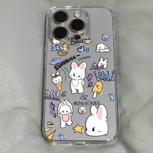 May include: A clear phone case with a variety of colorful cartoon stickers. The stickers feature bunnies, ice cream cones, stars, and other whimsical designs. The text "SURPRISE" and "LEE KNOW" are visible on the case.