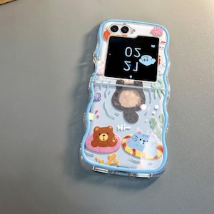 May include: A blue and white phone case with a cartoon bear, cat, and otter design. The case has a small screen on the front that displays the time, 1:50.