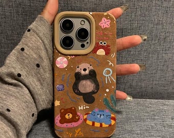 May include: A brown phone case with a painted design of a sea otter, a crab, a starfish, a jellyfish, and two animals in inner tubes. The text "Hi" is written on the case.