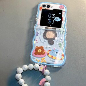 May include: A blue phone case with a cartoon design of a bear, cat, and otter swimming. The case has a clear window on the front that shows a digital clock displaying "15:30".