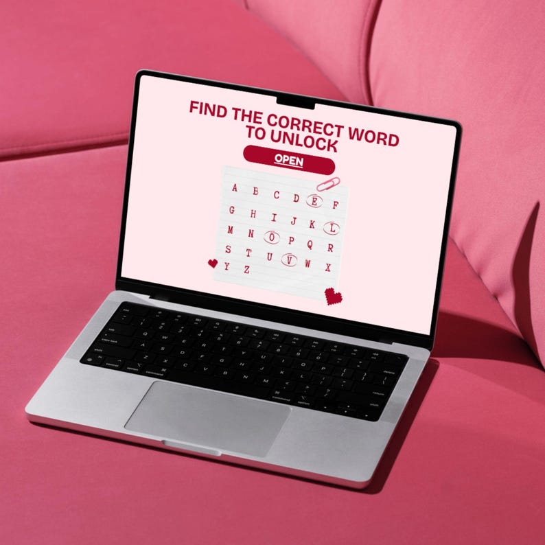 May include: A silver laptop displaying a puzzle game with the text "FIND THE CORRECT WORD TO UNLOCK". The game features a grid of letters and the word "OPEN". The laptop is on a pink surface.