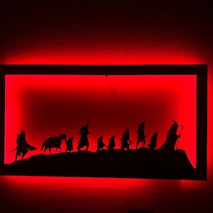 Può includere: Una silhouette nera in metallo della Compagnia dell'Anello della serie di film Il Signore degli Anelli, montata in una cornice rettangolare con luci LED rosse.