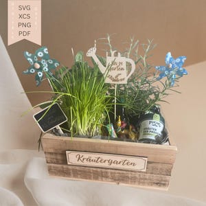 May include: A wooden planter box filled with herbs, decorative pinwheels, and signs. The box has the word "Kräutergarten" on the front. A sign reads "BIN IM Garten". A bottle of "FUCHS Kräuter der Provence" is visible.