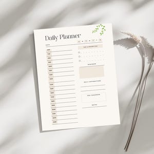 Könnte beinhalten: Ein cremefarbener Tagesplaner mit dem Titel "Daily Planner" in einer minimalistischen Schriftart. Der Planer enthält Abschnitte für Zeitfenster, oberste Prioritäten, Erinnerungen, tägliche Affirmationen und Notizen. Ein Zweig grüner Blätter verziert die obere rechte Ecke.