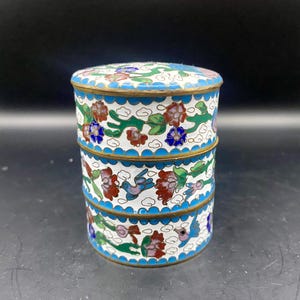 May include: A three-tiered, round trinket box with a white background and floral designs in red, blue, and green. The box has a blue scalloped border and gold trim. The lid also features a floral pattern.