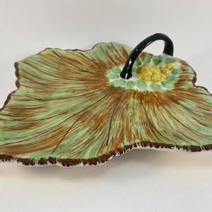 May include: A green and brown ceramic leaf-shaped serving dish with a black handle. The dish has a raised flower design in the center with yellow and green accents.