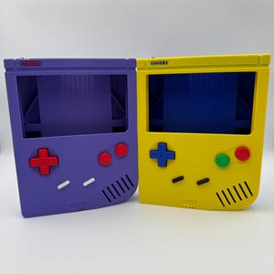 Puede incluir: Dos contenedores de almacenamiento coloridos con forma de consolas de juegos portátiles retro. Uno es morado con botones rojos, el otro es amarillo con botones azules y verdes. Los textos "CONDER" y "GONORA" son visibles.