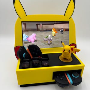 May include: A yellow and black Pikachu-themed gaming console with a screen displaying a video game. The console features a Pikachu figure, game cartridges, and controllers. The design includes Pikachu ears.