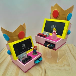 May include: Two pink Nintendo Switch console stands featuring a yellow screen frame and a large gold crown topper with blue and red accents. Each stand holds a console, game cartridges, a controller, and a small character figurine of Mario or Peach.