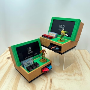 May include: Two tan and green 3D-printed Nintendo Switch storage organizers featuring engraved circular patterns. Each unit holds a console screen, game cartridges, and a controller, with a small figurine of Link or Samus standing on a circular base.