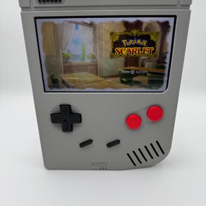 以下が含まれることがあります： 「Pokémon Scarlet」のシーンが表示された画面を持つ、グレーの携帯ゲーム機。 方向パッド、赤いボタン、そして「PRESS A BUTTON」の文字があります。 コンソールの上面には「COHOBA」の文字があります。