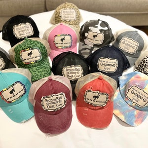 May include: A collection of trucker hats with various designs and colors. The hats feature a mesh back and a distressed look. Some hats have the text "Bronco Hair Don't Care" or "Girl". Other hats have a "Bronco" logo.