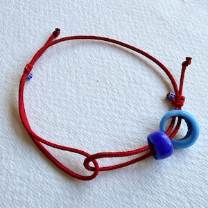 Könnte beinhalten: Rotes Kordelarmband mit blauen und lila Perlen. Das Armband hat einen Schiebeknotenverschluss für verstellbare Größe. Die Perlen sind eine Mischung aus runden und zylindrischen Formen und setzen Farbakzente.