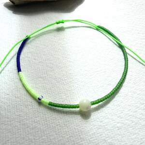 May include: A beaded necklace on a neon green cord featuring a sequence of dark blue, light green, and forest green seed beads. The design includes a single small white bead with a blue stripe and a larger, smooth, off-white spherical bead.