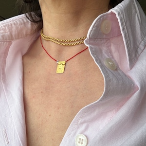 May include: A person wearing a pink button-down shirt with a gold chain necklace and a red string necklace with a small gold pendant.