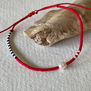 Pode incluir: Colar de contas vermelho com uma pérola branca e cordão vermelho ajustável. O colar apresenta contas listradas em preto e branco e listras brancas. A joia é exibida em uma superfície branca.