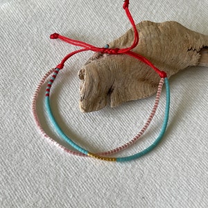 May include: A layered bracelet with pink, blue and gold beads. The bracelet is tied with a red string.