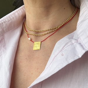 May include: A gold chain necklace with a small gold pendant and a red string necklace with a white pearl charm. The necklaces are layered on a person's neck.
