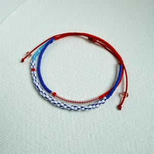 May include: A double-strand bracelet featuring a red cord with adjustable knots and two rows of beads. One row consists of white beads with blue stripes, while the other row features a mix of light blue, dark blue, and translucent pink beads.