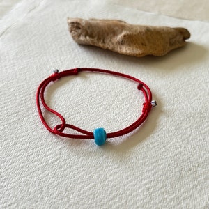 Puede incluir: Un brazalete de cuerda roja con una cuenta azul. El brazalete está atado con un nudo y tiene una pequeña cuenta blanca en cada extremo.