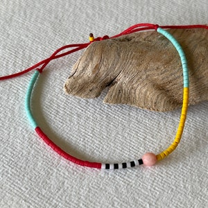 May include: A colourful beaded necklace with a red cord. The necklace features sections of red, light blue, yellow, and black and white beads, with a pink bead accent. The necklace is displayed on a piece of driftwood.