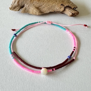 Puede incluir: Dos pulseras de cuentas. Una pulsera es turquesa y rosa, la otra es rosa y marrón. Una cuenta blanca está centrada en las pulseras. Las pulseras están sobre una superficie blanca con un trozo de madera flotante en el fondo.