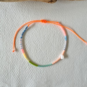 Könnte beinhalten: Ein farbenfrohes Perlenarmband mit einem weißen Perlenanhänger. Das Armband besteht aus kleinen, bunten Perlen in Blautönen, Grün, Rosa und Orange. Das Armband ist mit einer leuchtend orangenen Schnur gebunden.