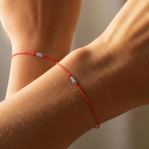 Può includere: Due braccialetti di filo rosso, ciascuno con un piccolo accento di perline color argento. I braccialetti sono indossati sui polsi, suggerendo un tema di amicizia o di coppia. Lo sfondo è di un tono morbido e neutro.