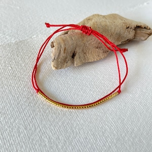 May include: A red string bracelet with a gold bead accent. The bracelet is tied in a knot and is ready to be worn.