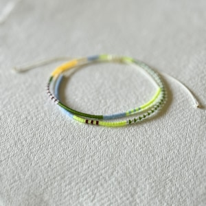 Puede incluir: Una delicada pulsera de cuentas con una mezcla de colores, incluyendo amarillo, verde, azul y granate. La pulsera está hecha de pequeñas cuentas coloridas ensartadas en un fino cordón blanco. La pulsera se muestra sobre una superficie texturizada blanquecina.