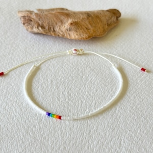May include: White beaded bracelet with a rainbow of colored beads. The bracelet is tied with white string and has red beads at the ends.