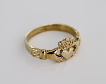 May include: A gold-colored Claddagh ring. The ring features a heart held by two hands, topped with a crown. The ring is set against a white background.