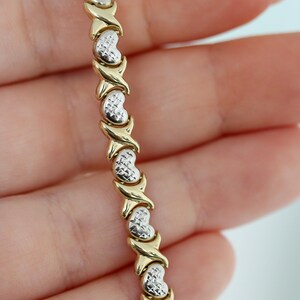 Puede incluir: Una pulsera de oro y plata con un patr&oacute;n repetido de eslabones en forma de coraz&oacute;n y conectores en forma de X. La pulsera se sostiene en una mano, mostrando el contraste entre los elementos de oro y plata.