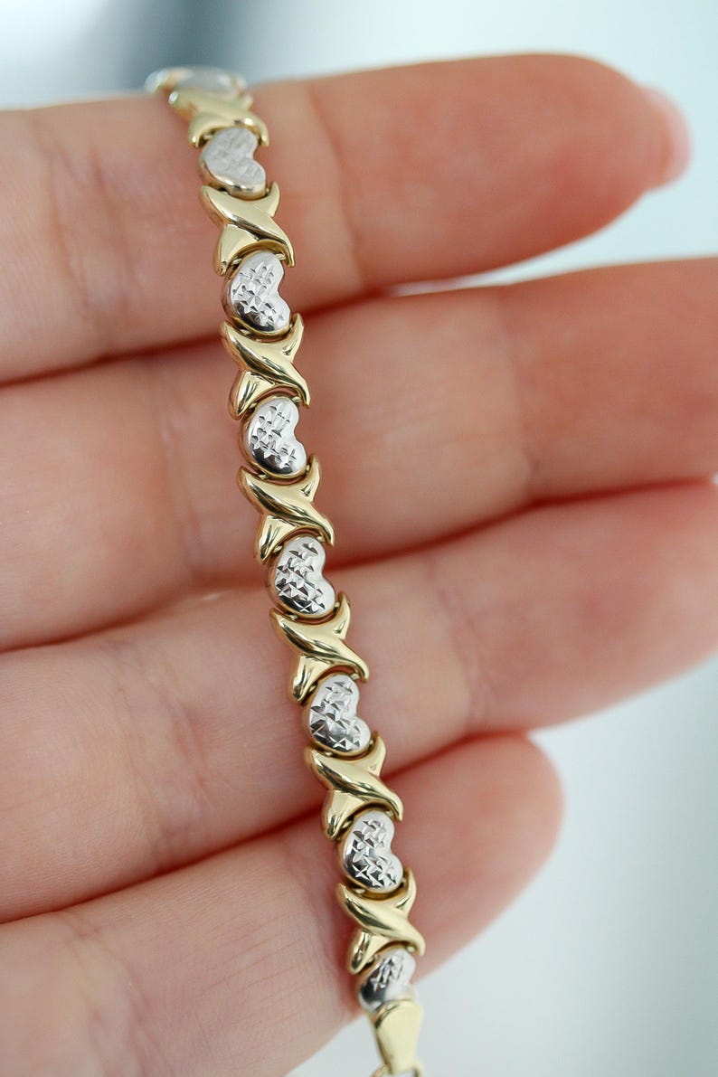 Puede incluir: Una pulsera de oro y plata con un patr&oacute;n repetido de eslabones en forma de coraz&oacute;n y conectores en forma de X. La pulsera se sostiene en una mano, mostrando el contraste entre los elementos de oro y plata.
