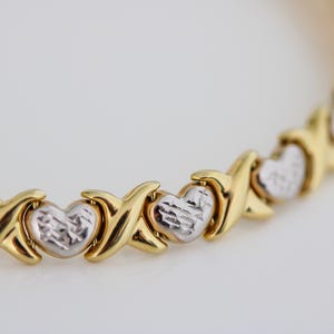 Puede incluir: Pulsera dorada con eslabones en forma de X y coraz&oacute;n alternados. Los corazones tienen un acabado plateado texturizado, mientras que los eslabones en X son de oro pulido. La joya se muestra sobre un fondo blanco.