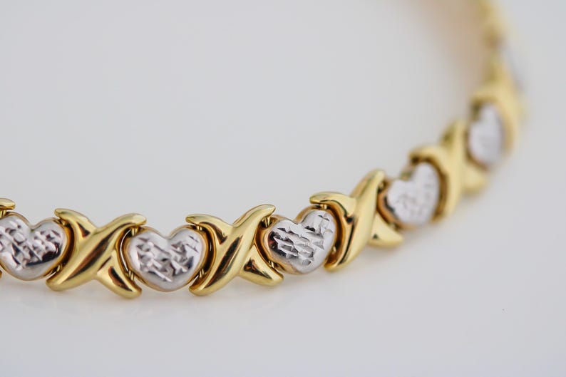 Puede incluir: Pulsera dorada con eslabones en forma de X y coraz&oacute;n alternados. Los corazones tienen un acabado plateado texturizado, mientras que los eslabones en X son de oro pulido. La joya se muestra sobre un fondo blanco.