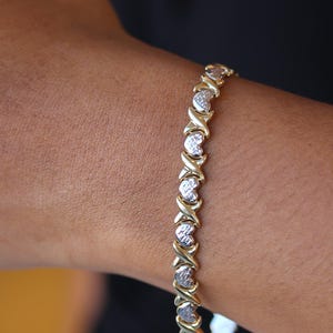 Puede incluir: Pulsera de oro y plata con un patr&oacute;n repetido de X y corazones. La pulsera se lleva en la mu&ntilde;eca, mostrando las X doradas y los eslabones plateados en forma de coraz&oacute;n. La joya es de dise&ntilde;o cl&aacute;sico.