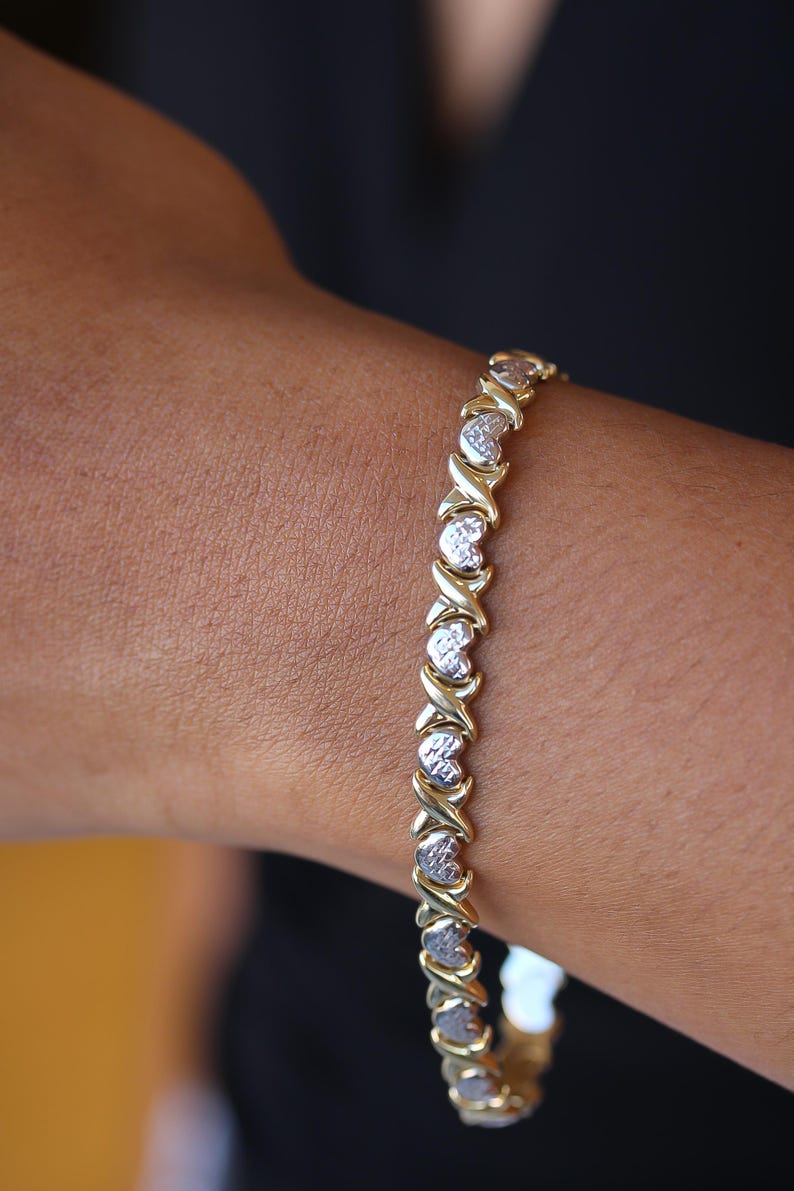 Puede incluir: Pulsera de oro y plata con un patr&oacute;n repetido de X y corazones. La pulsera se lleva en la mu&ntilde;eca, mostrando las X doradas y los eslabones plateados en forma de coraz&oacute;n. La joya es de dise&ntilde;o cl&aacute;sico.