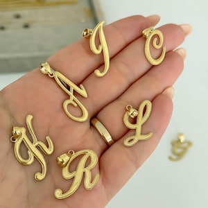 May include: Gold-colored initial pendants in a variety of cursive fonts. The pendants are displayed on a hand, showcasing the letter designs. The pendants are likely made of gold and are designed for necklaces or bracelets.