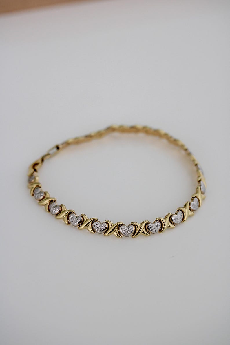 Puede incluir: Pulsera dorada con un patr&oacute;n repetido de corazones y X. La pulsera tiene un dise&ntilde;o de cadena delicado y probablemente est&aacute; hecha de oro o metal chapado en oro. Los corazones tienen una apariencia texturizada.
